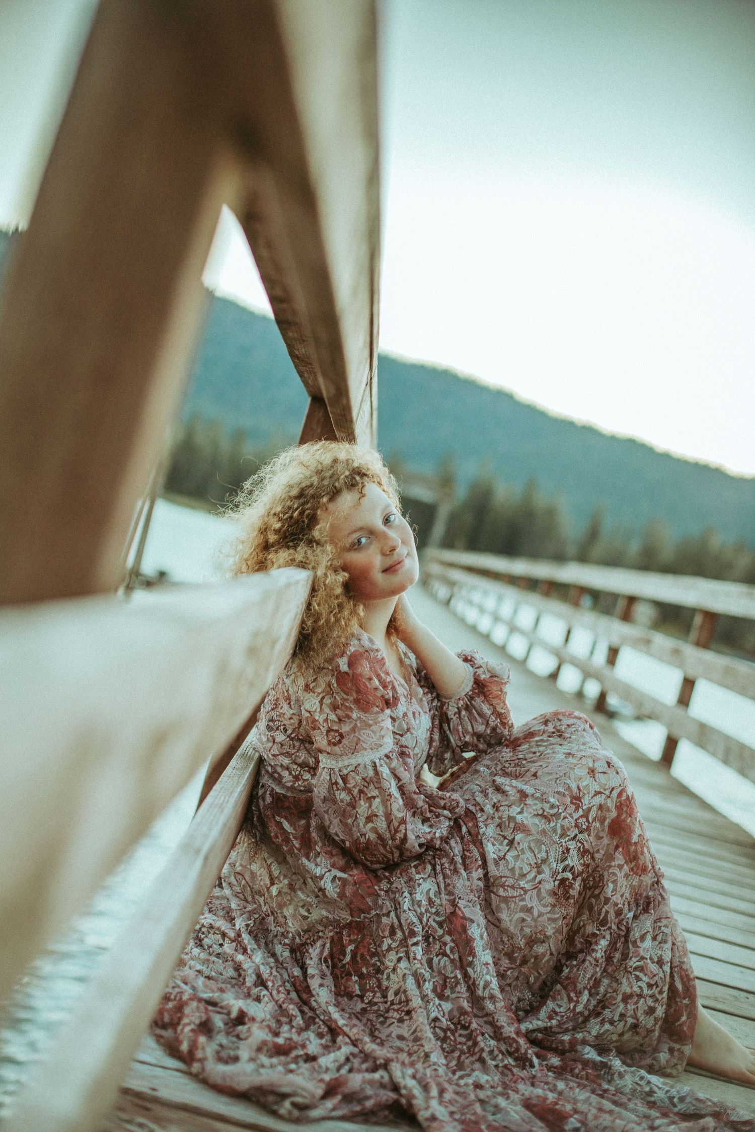 A dreamy portrait in a floral vintage dress sitting on a wooden deck with mountains in the background.