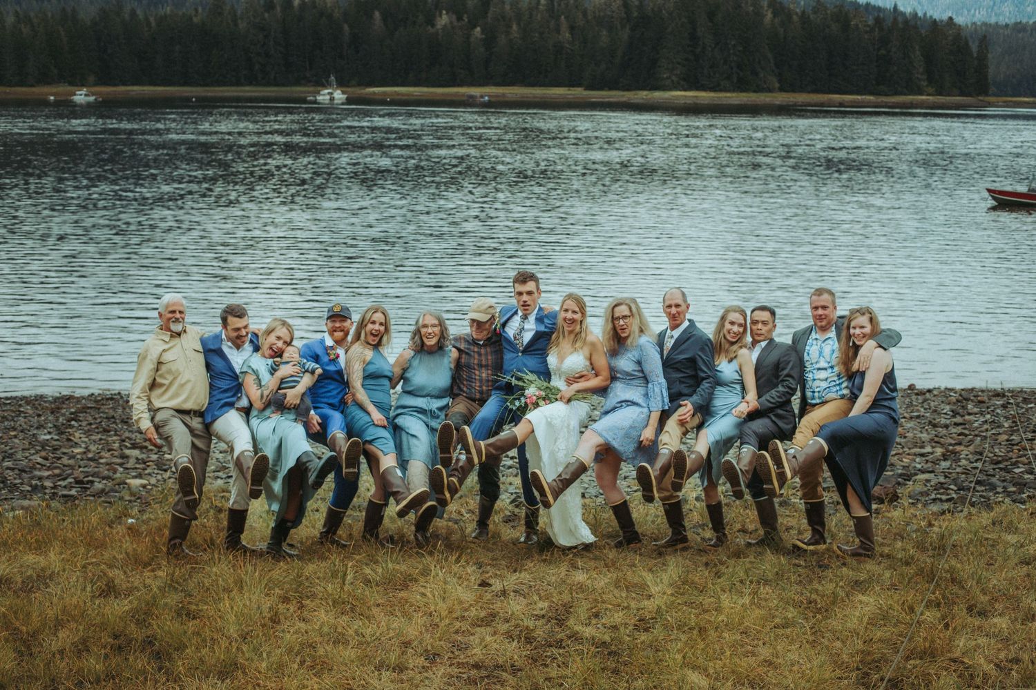Large wedding party in blue and white attire poses casually by a scenic mountain lake in Alaska.
