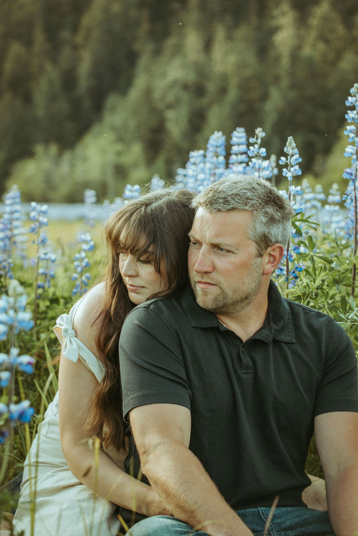 Engaged Couple sits close together outdoors among blooming blue lupine flowers on a summer day in Southeast Alaska..