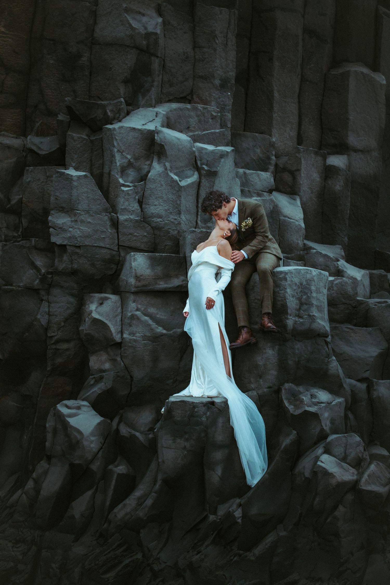Bride in white gown and groom in suit share a kiss on dramatic dark basalt rock columns in an outdoor wedding photo.