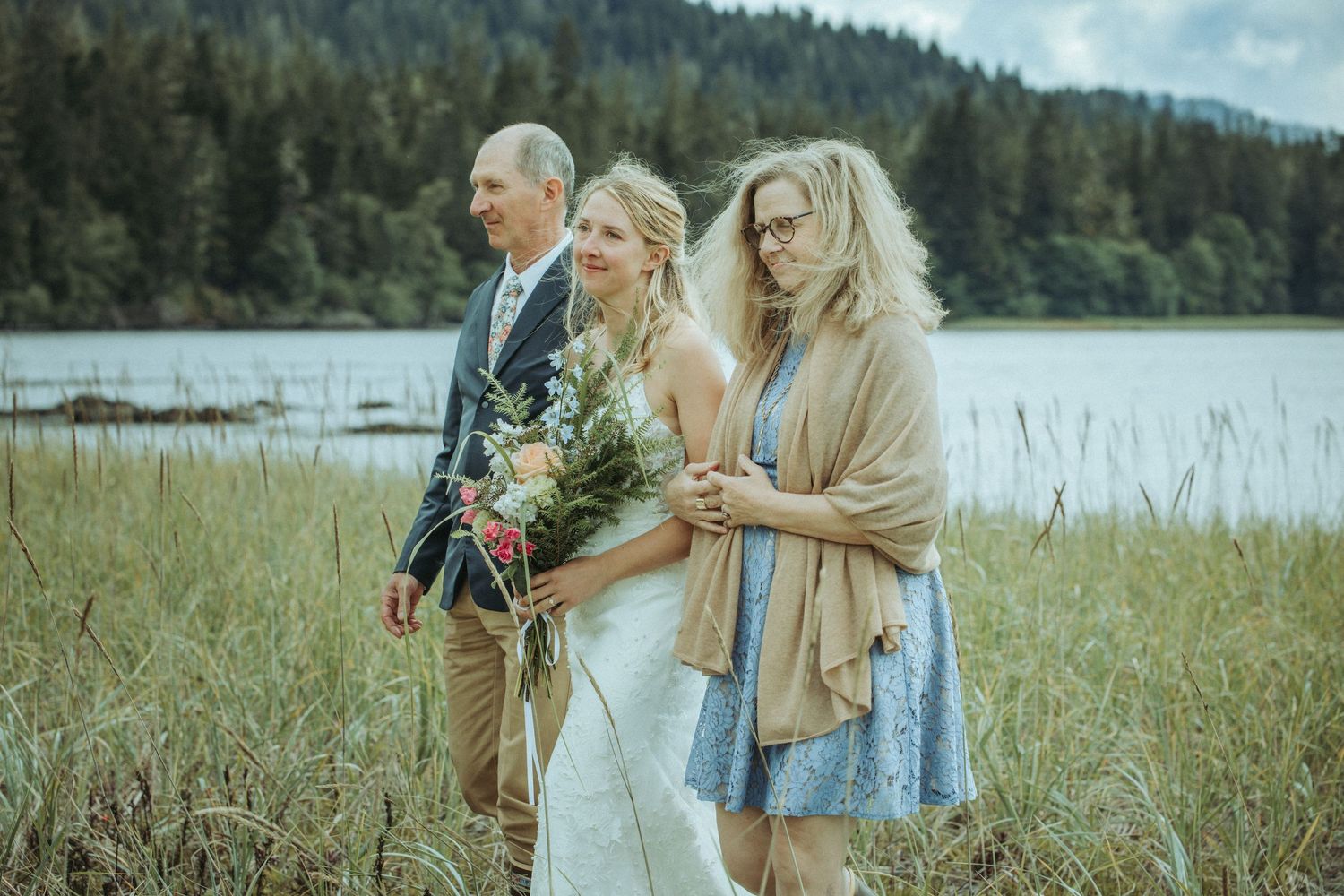 Wedding group stands in tall grass by lake with mountains and forest in background in Alaska.