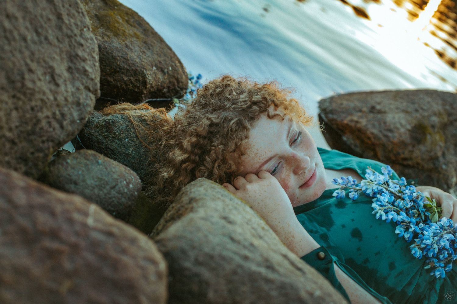 Artistic portrait against rocks with blue flowers and warm sunset lighting.