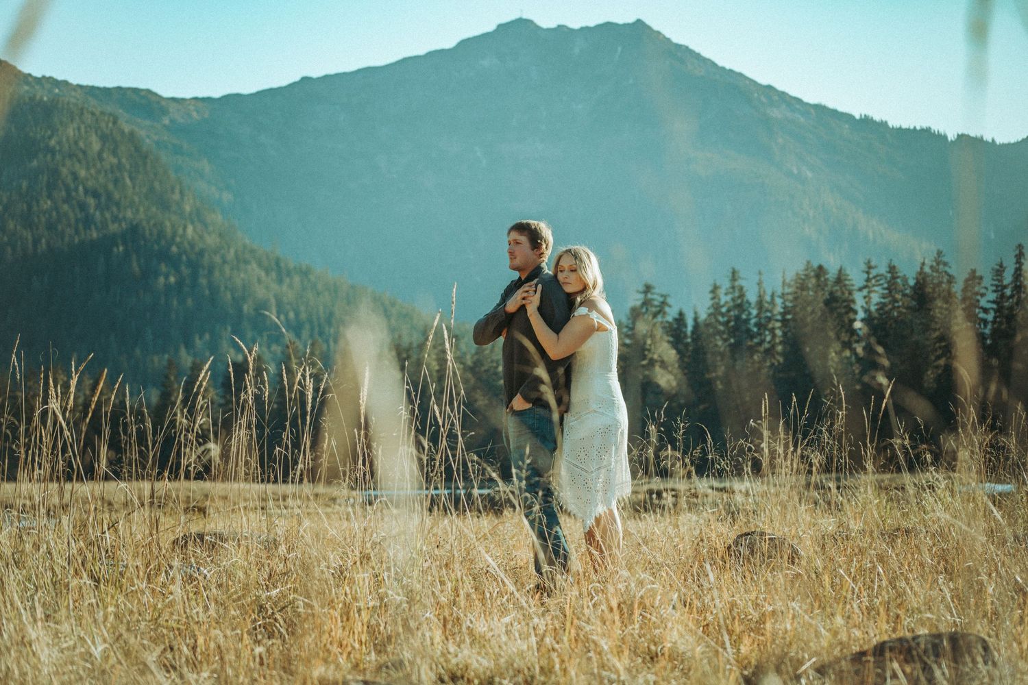 A couple embraces in a golden meadow with majestic mountains and evergreen forest in the background in Southeast Alaska