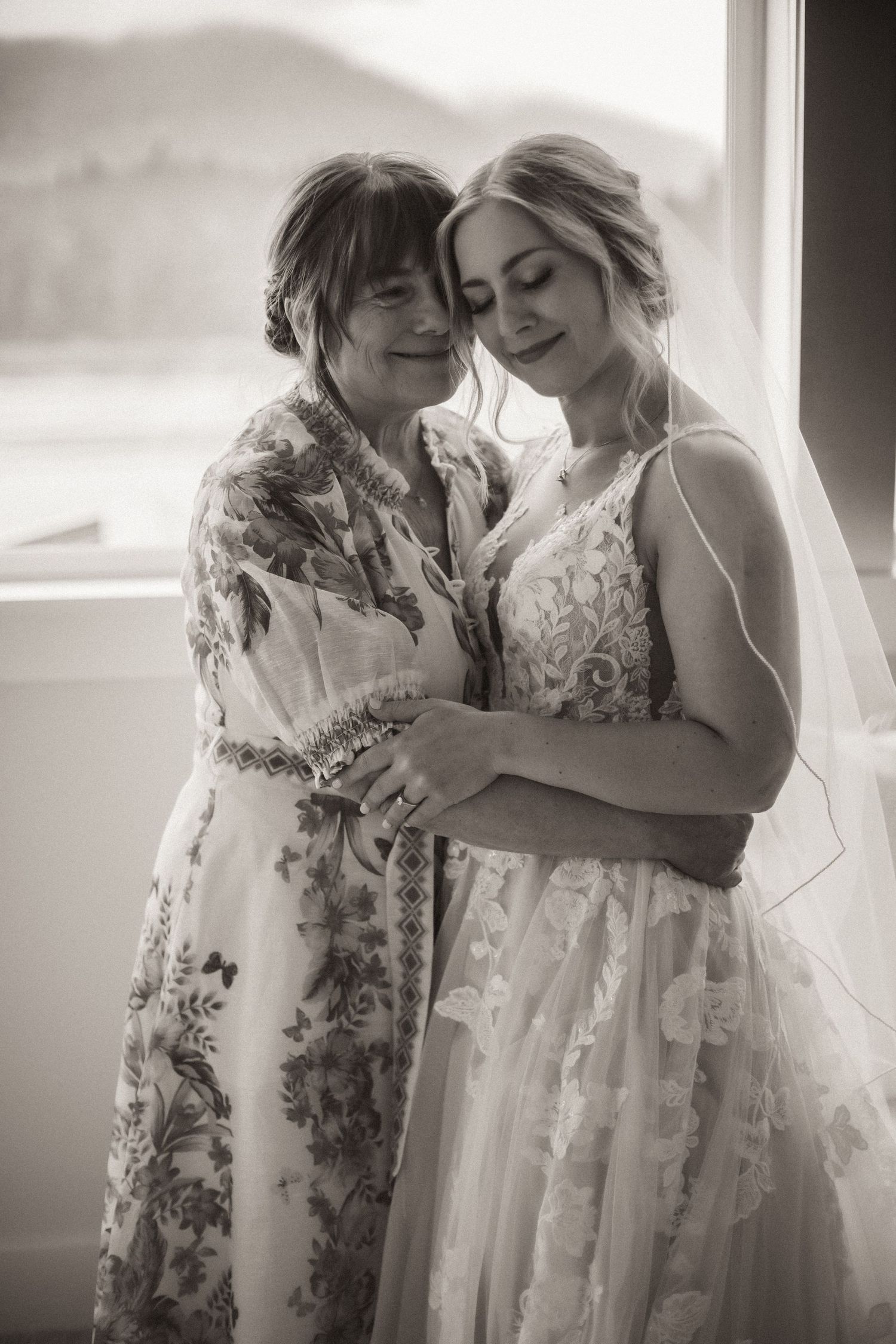 Black and white photo of a bride in lace gown and veil sharing a tender embrace with an older woman in a floral dress.