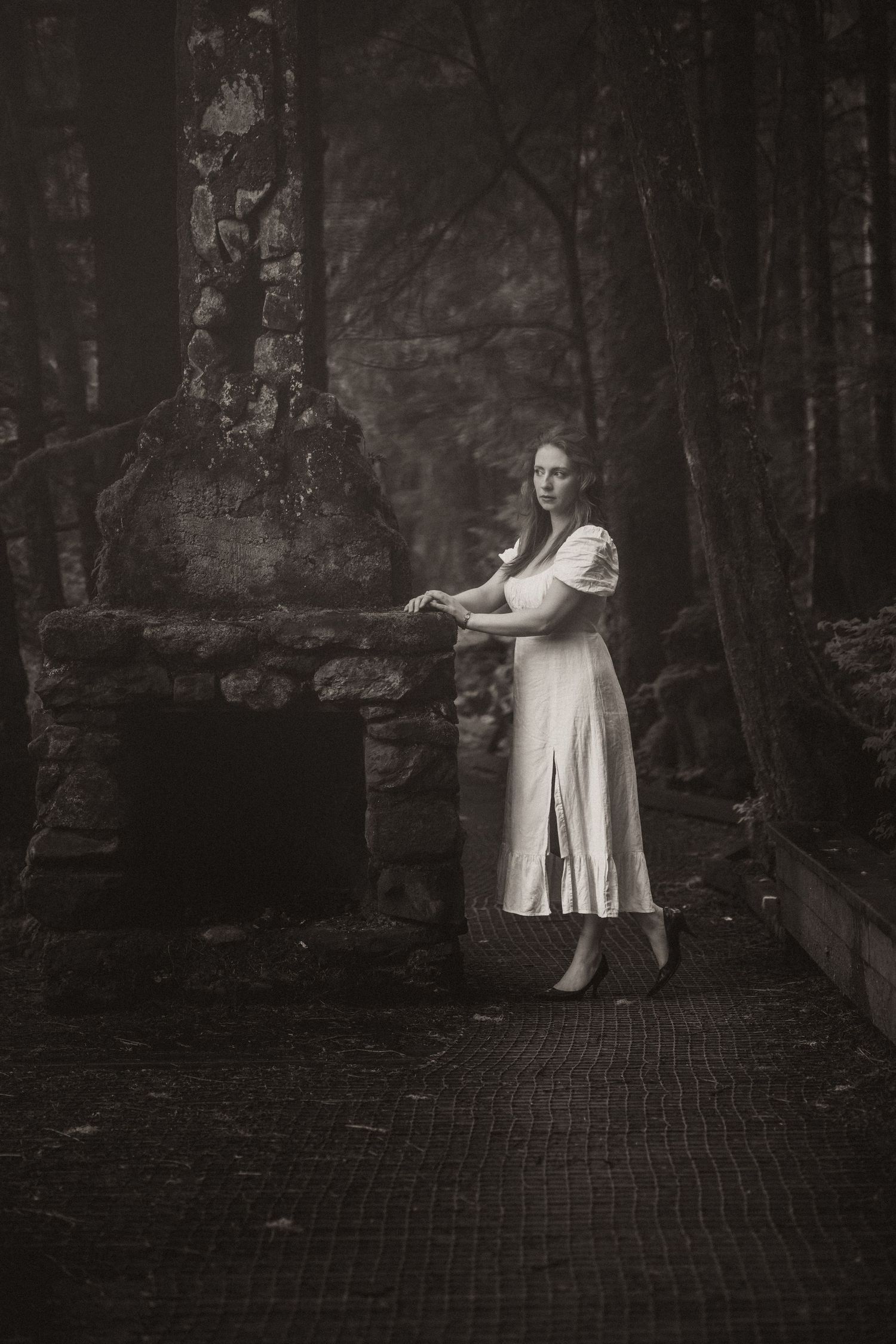 Woman in white dress standing by stone ruins in dark forest, black and white fine art photography.