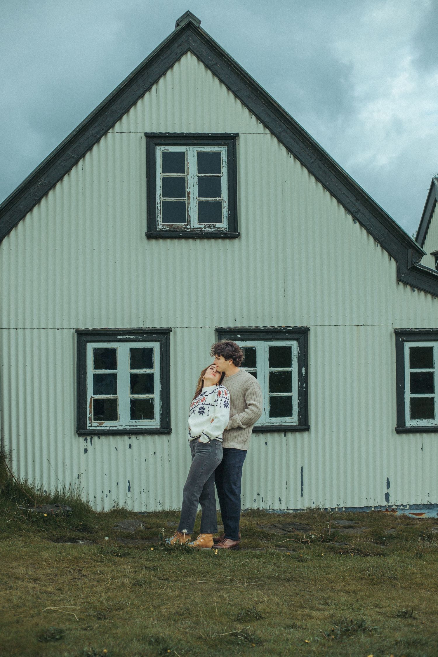 Couple embracing outside a white Scandinavian-style turf house with dark-trimmed windows while wearing cozy sweaters for Iceland Engagement.