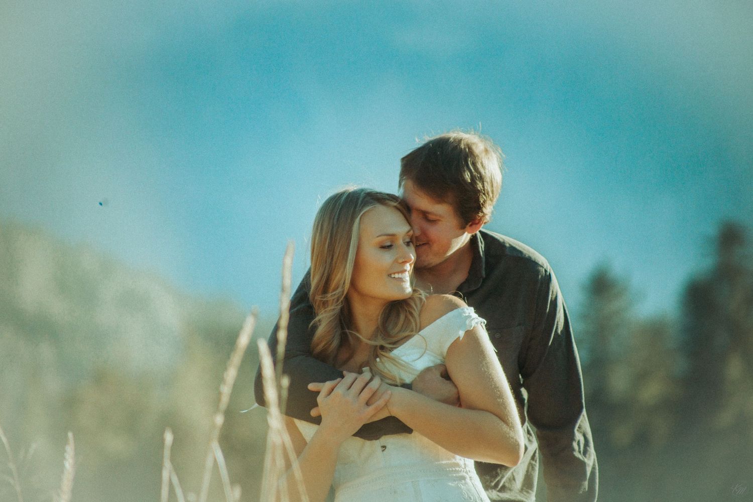 A couple embraces in a golden meadow with majestic mountains and evergreen forest in the background in Southeast Alaska