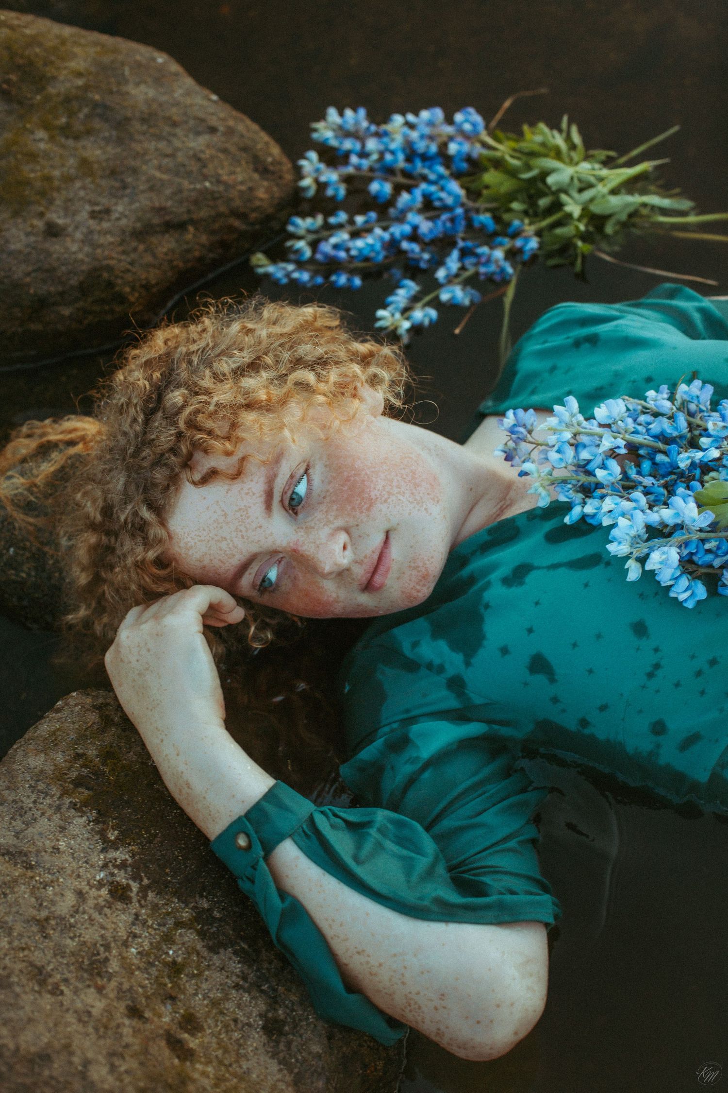 Dreamy lakeside portrait with blue flowers and teal dress against rocky shore.