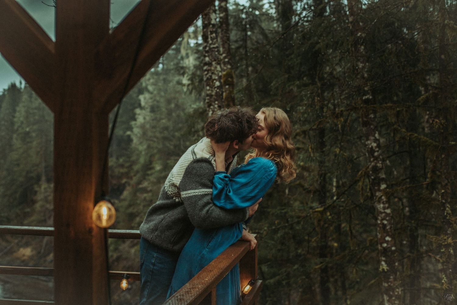A romantic couple embraces on a rustic wooden deck at dusk with moody forest scenery in the background in Southeast Alaska.