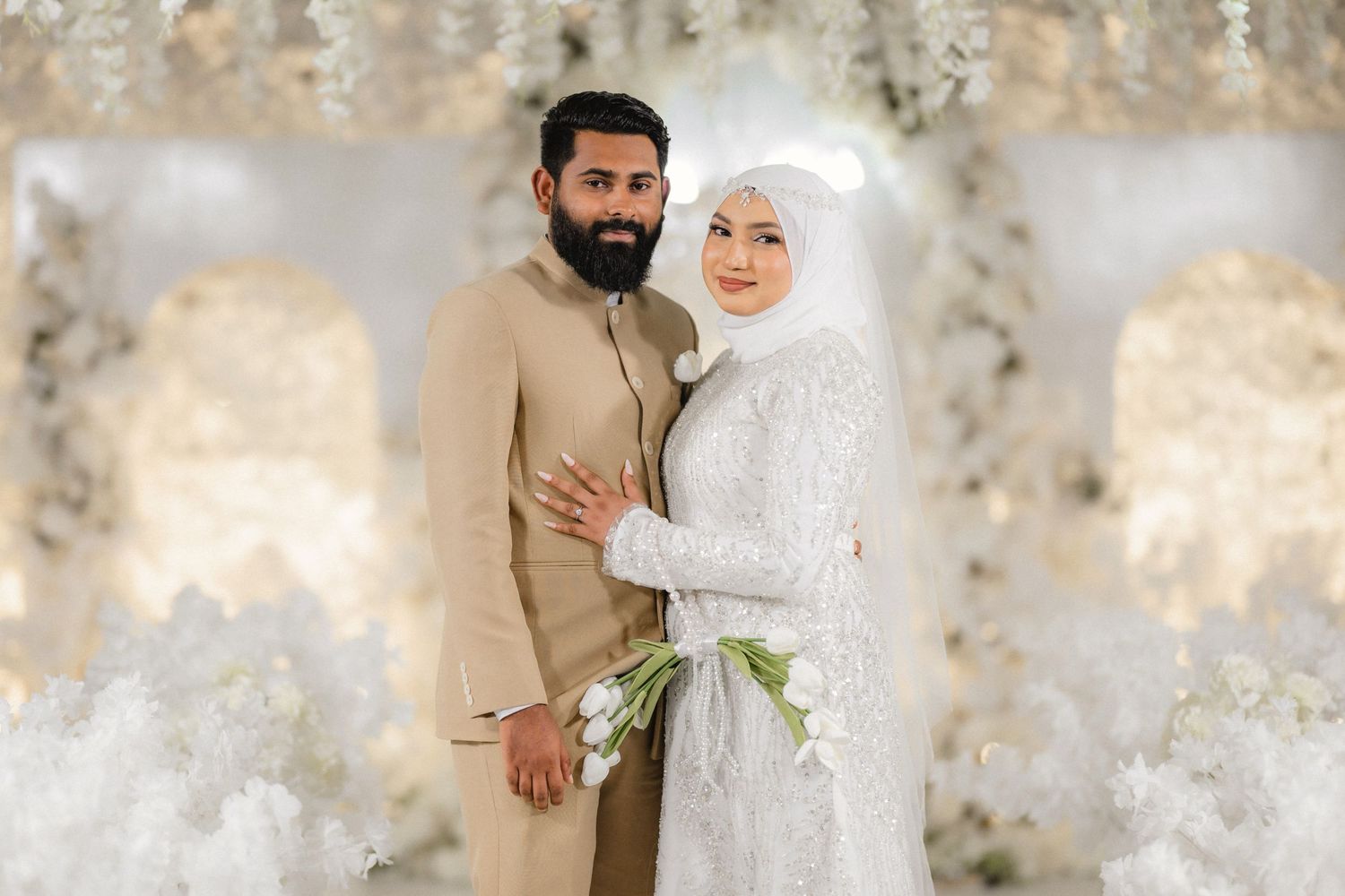 A couple in traditional Middle Eastern wedding attire pose together against a textured beige wall.