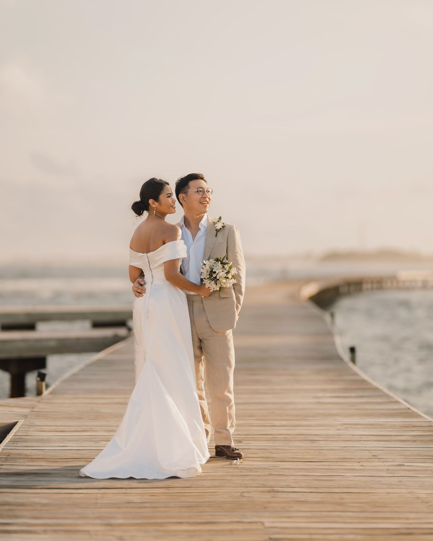 Couple in wedding attire standing on wooden dock by the ocean at sunset.