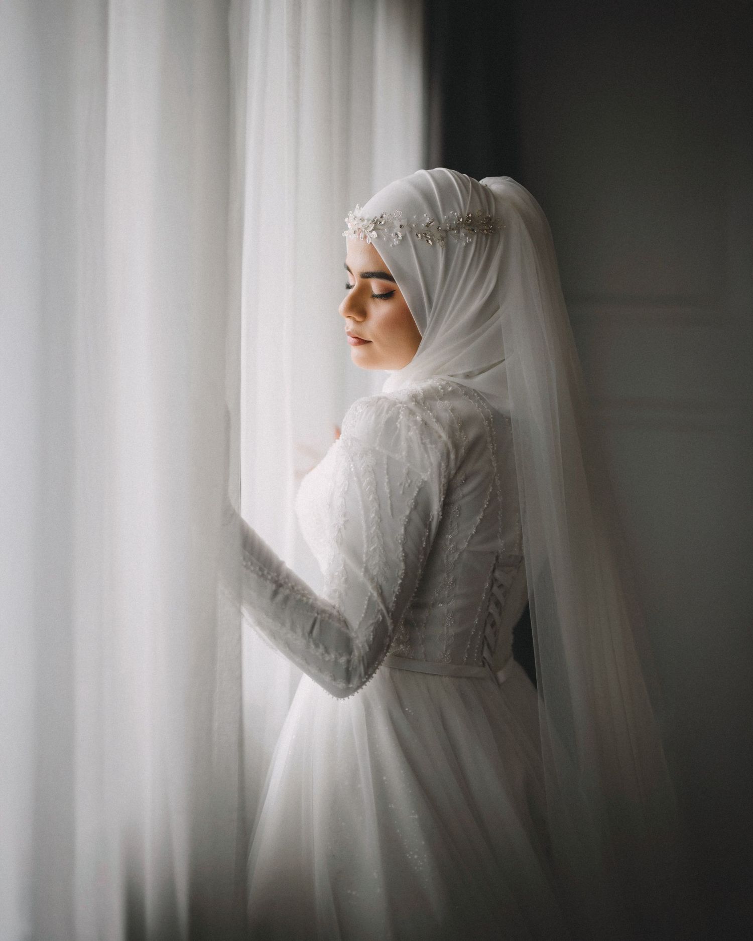 Bride in white lace dress and hijab poses gracefully by window with soft lighting.
