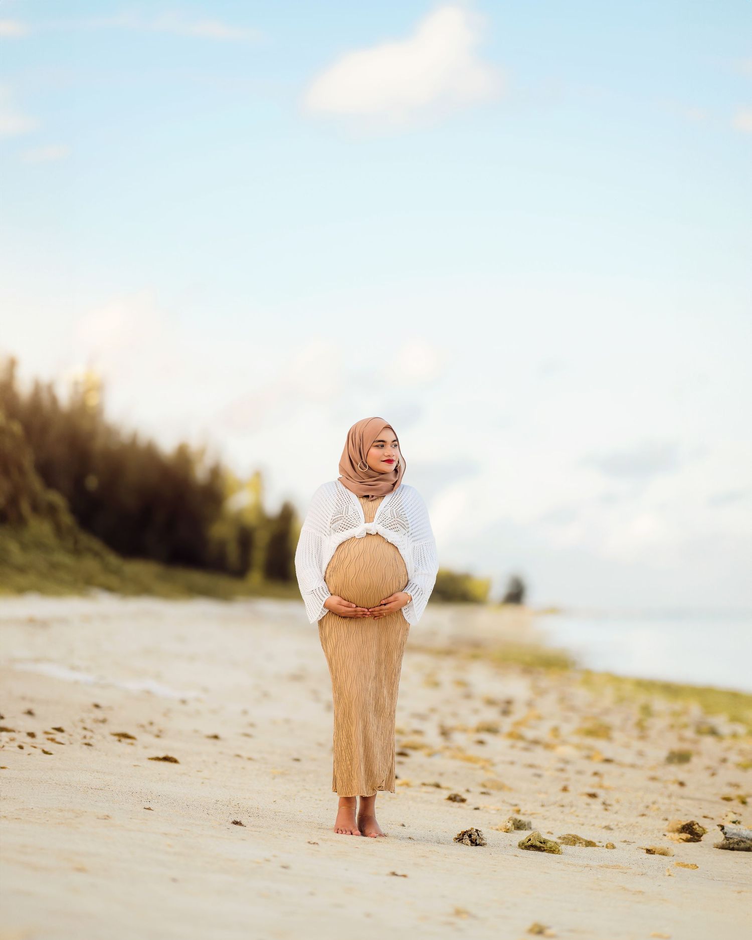 A person in a flowing beige dress stands on a sandy beach with ocean waves in the background.