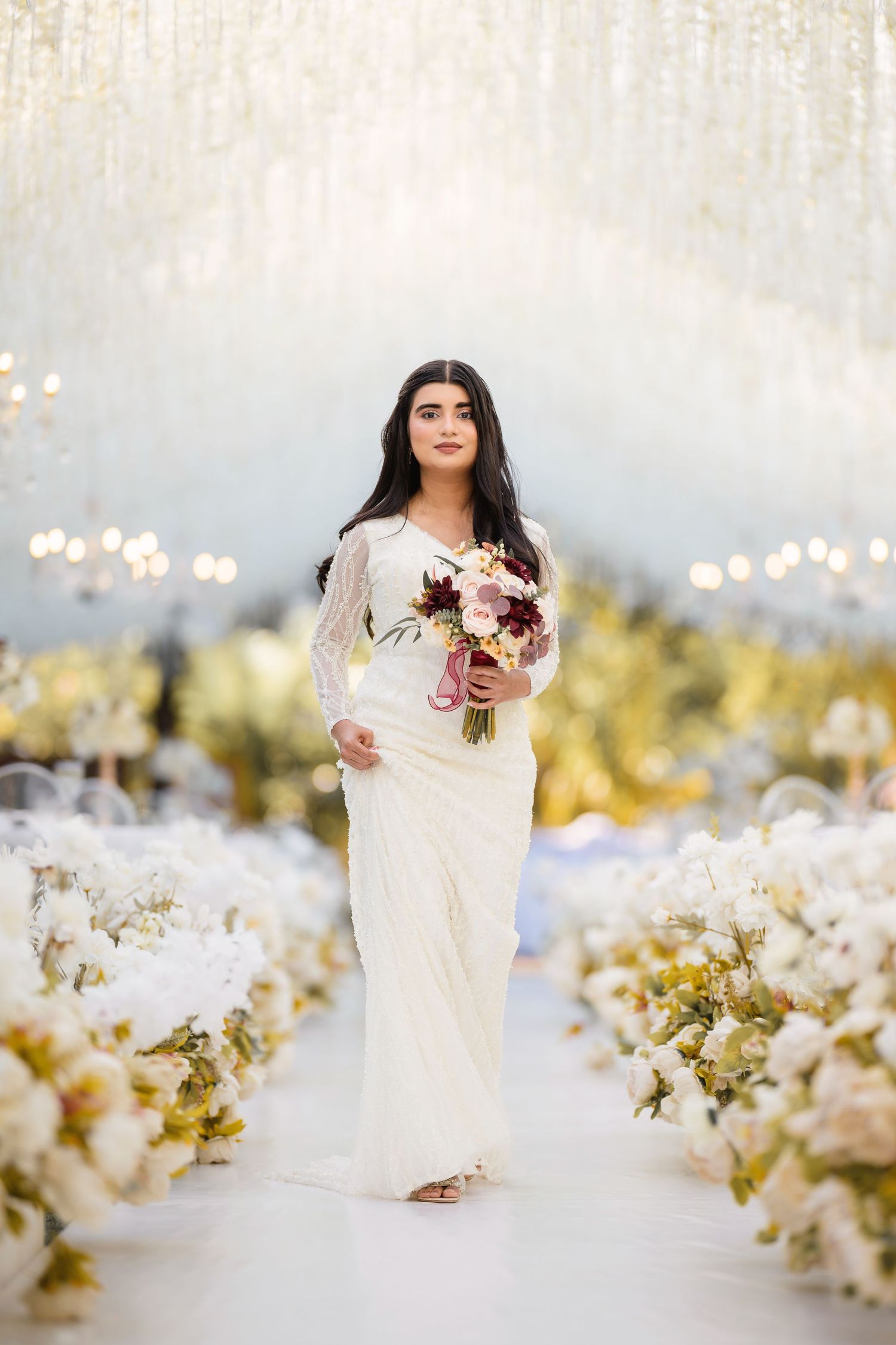 Bride in white dress poses elegantly against a dreamy backdrop with white floral decorations and soft bokeh lights.