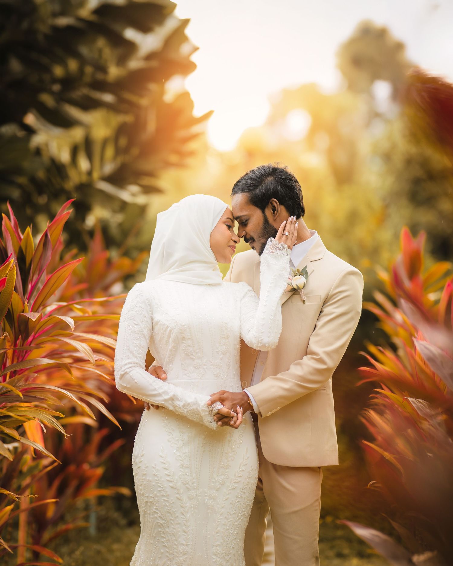 Couple shares tender moment among red tropical plants during sunset wedding photography.