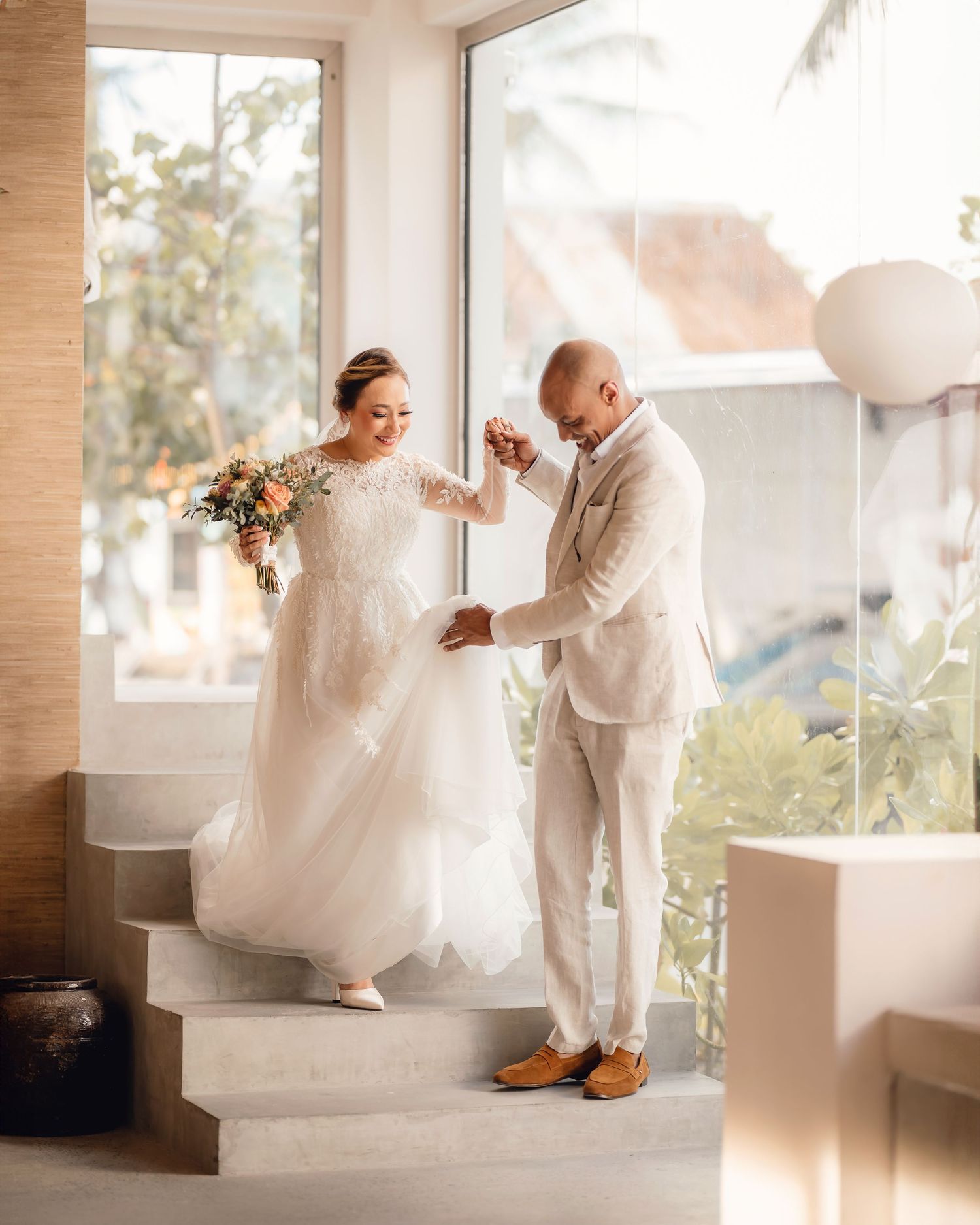 Playful wedding couple interacts on white stairs in sunlit indoor setting.