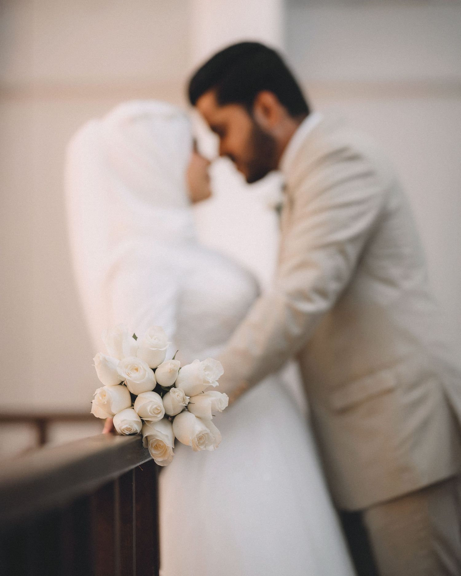 Wedding couple in white attire share tender moment with white rose bouquet.