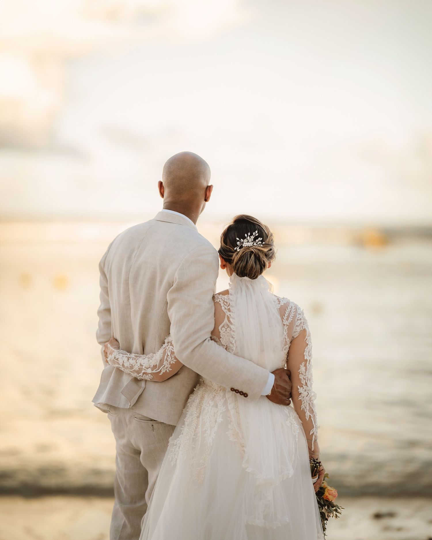 Newlywed couple embraces while standing on beach at sunset in romantic wedding photo.