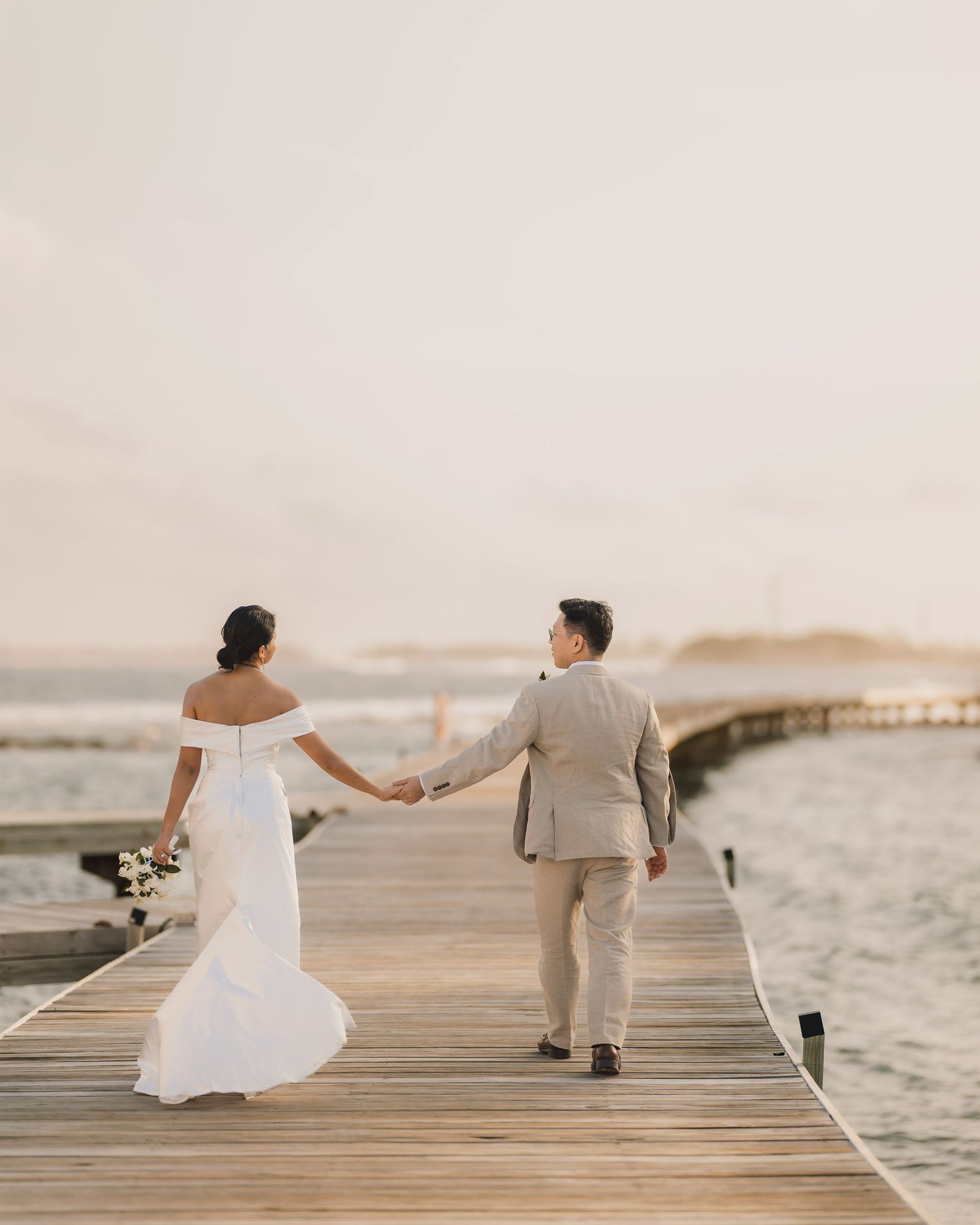 Wedding couple walking hand in hand on wooden pier by the ocean.
