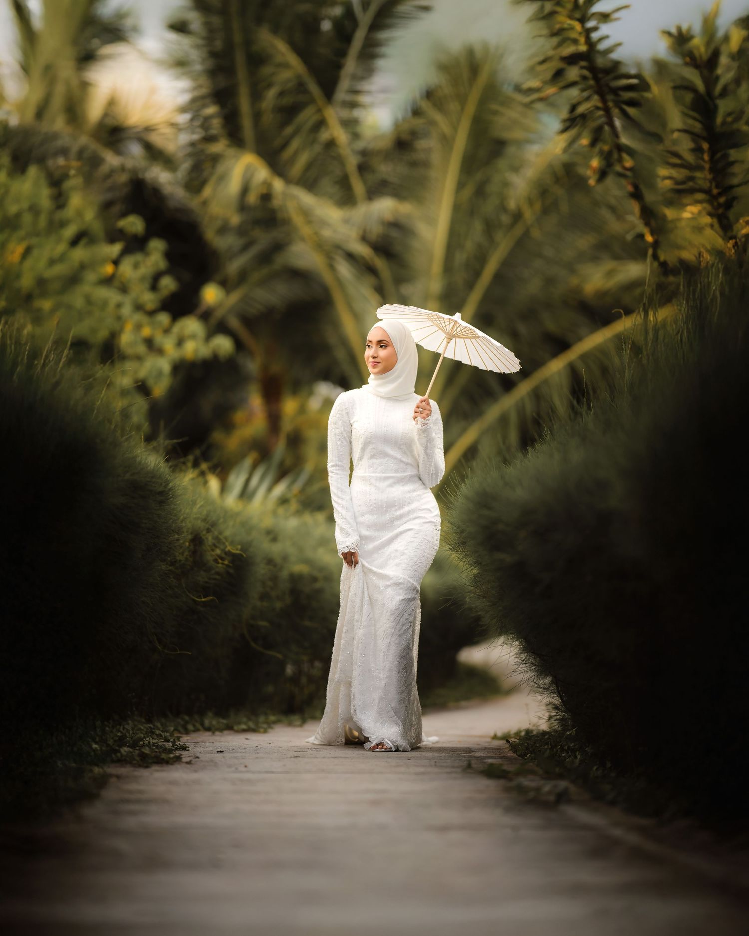 A bride in white dress and parasol walks along a palm-lined tropical pathway.