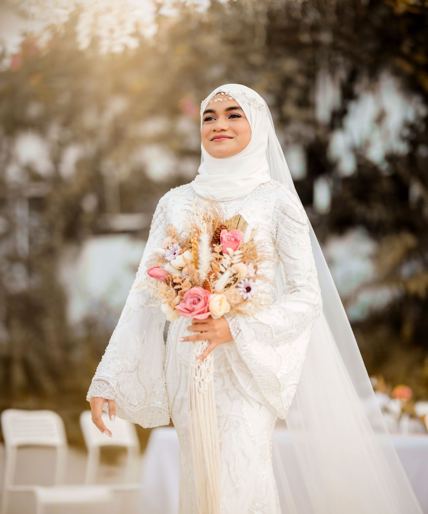 A bride in a white hijab and dress holds a pink and white bouquet against a soft outdoor background.