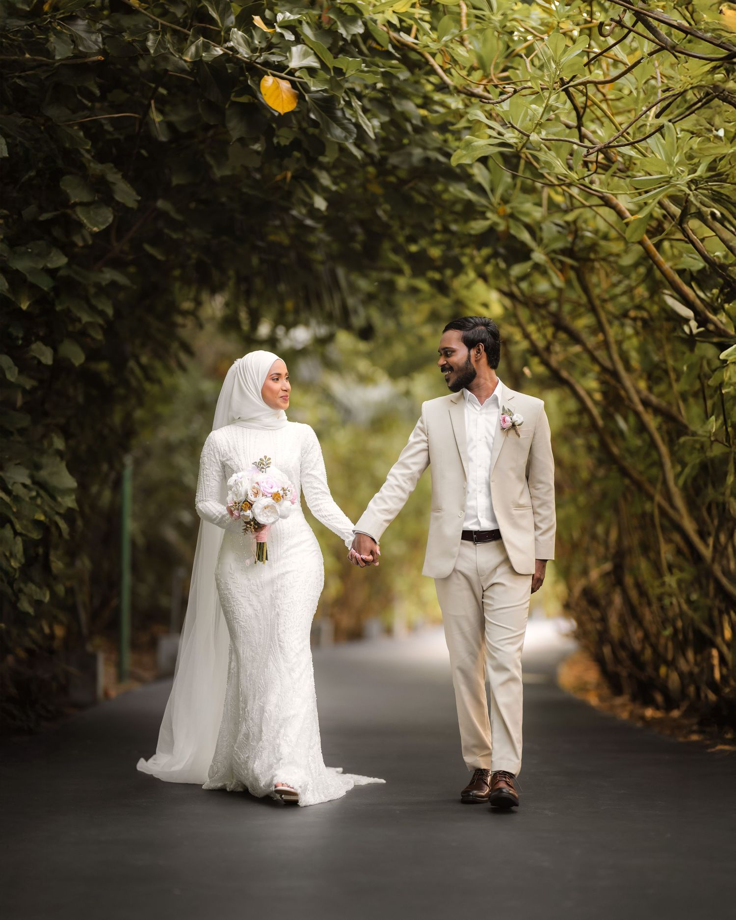 A couple in wedding attire walks along a tree-lined path during a romantic outdoor photo session.