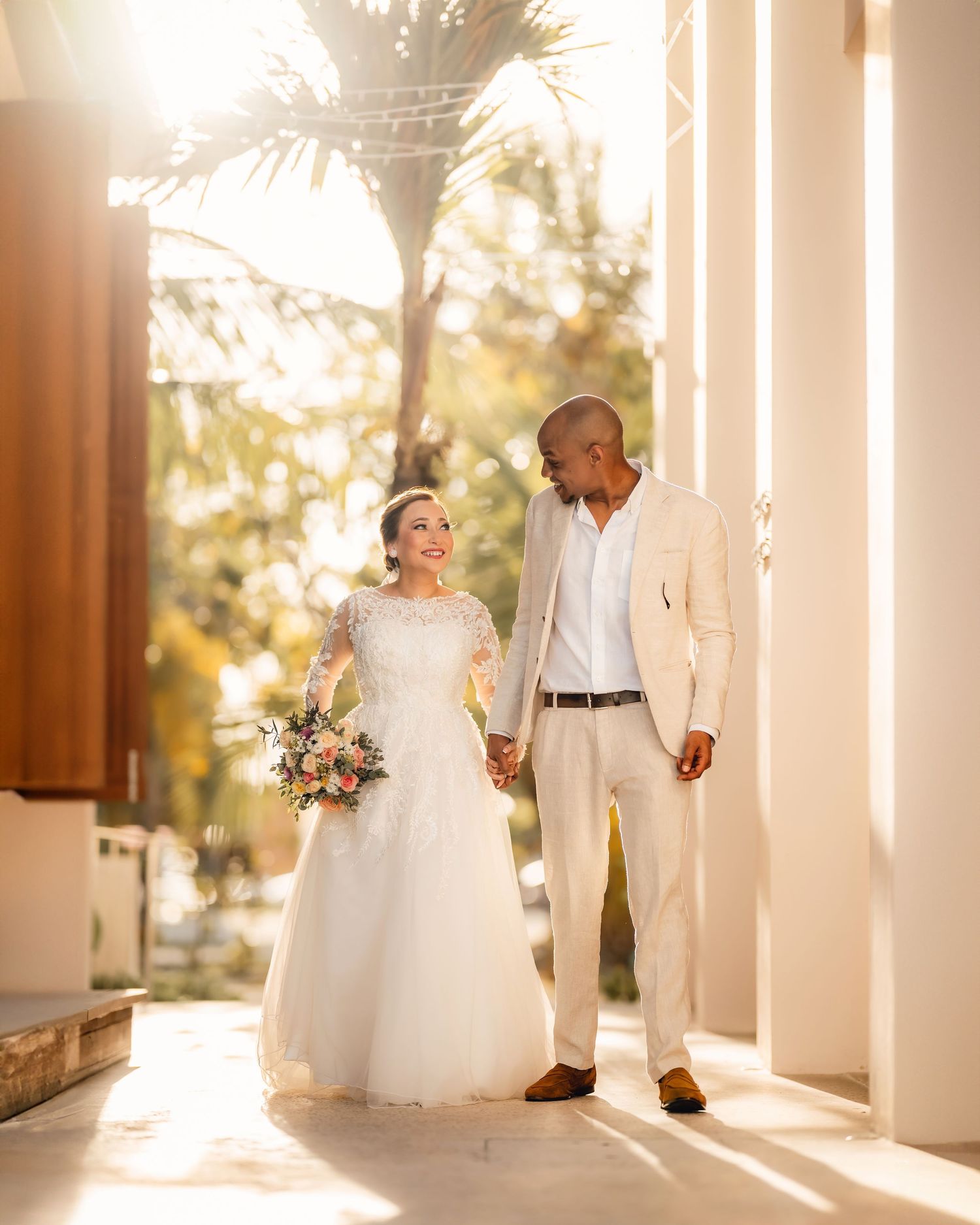 Wedding couple walks through sunlit colonnade creating romantic silhouettes against pillars.
