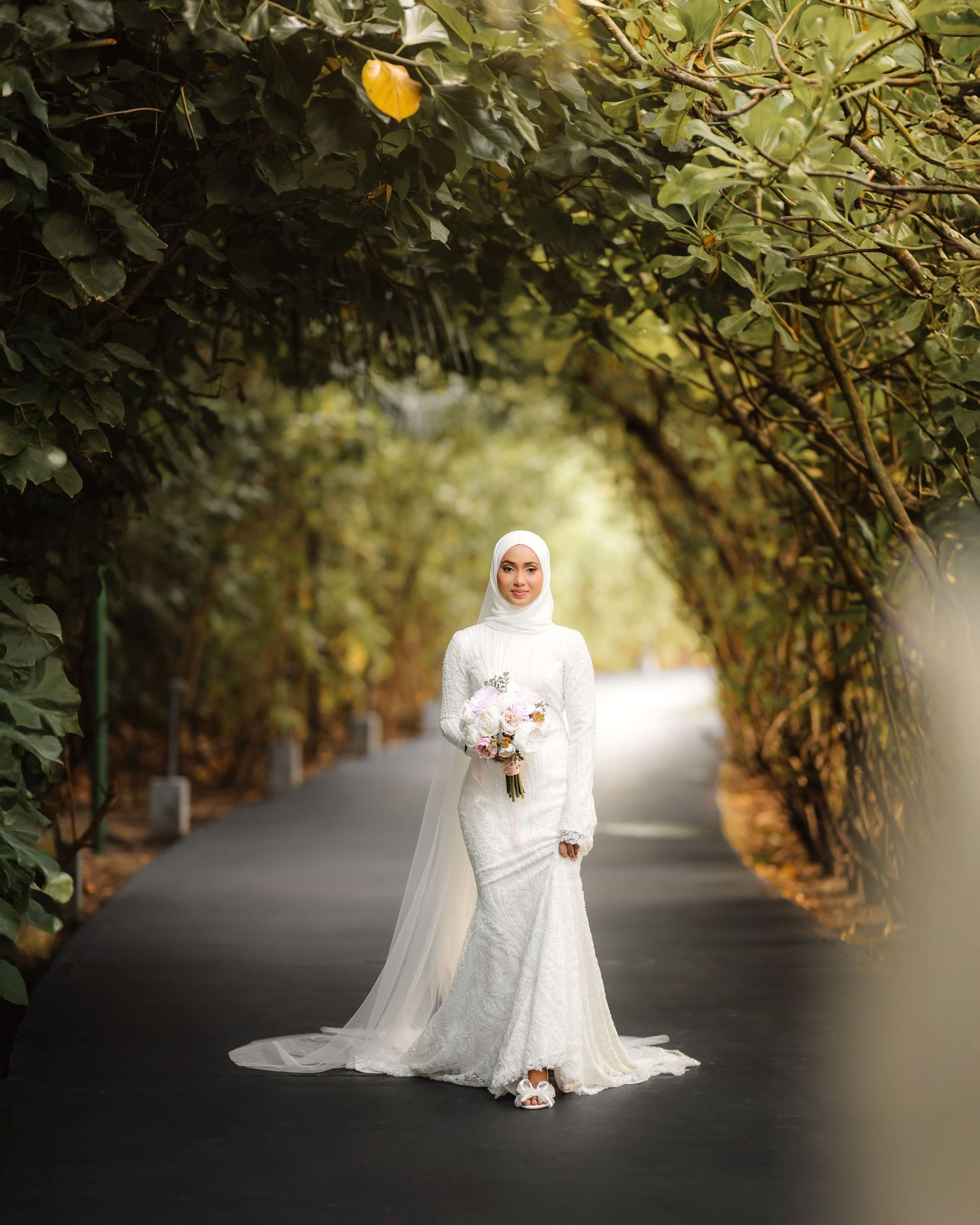 Bride in elegant white dress and hijab poses on a tree-lined pathway.