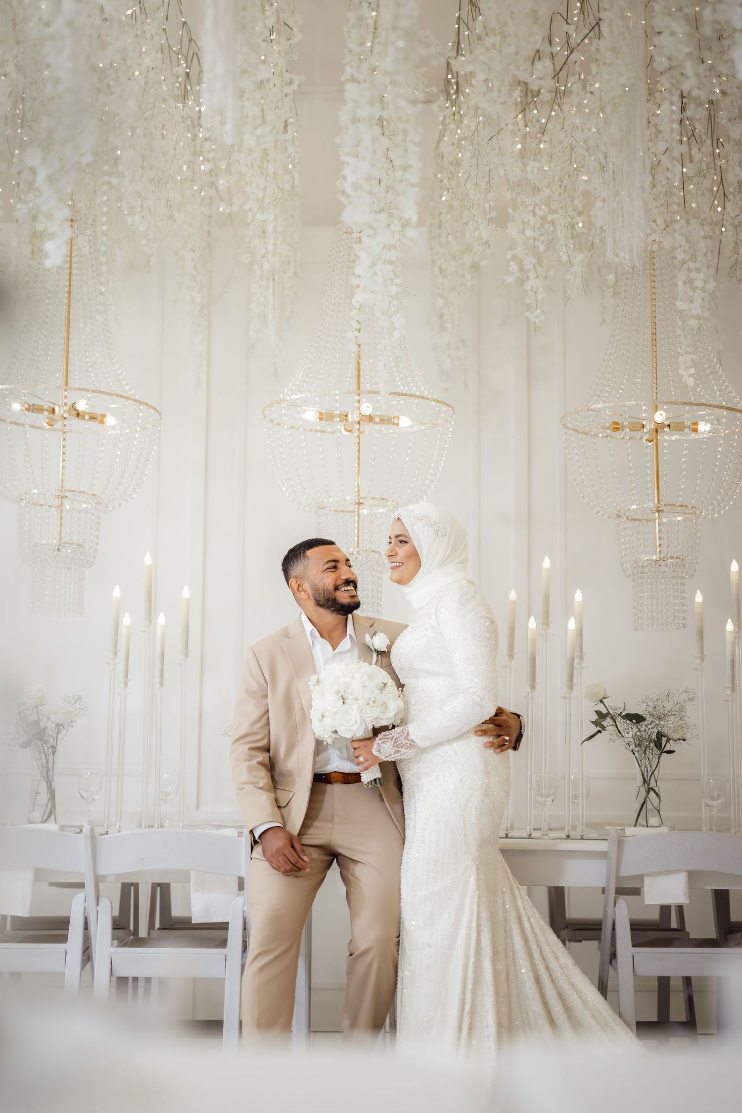 Elegant wedding couple poses in white decorated reception venue with hanging crystal chandeliers.