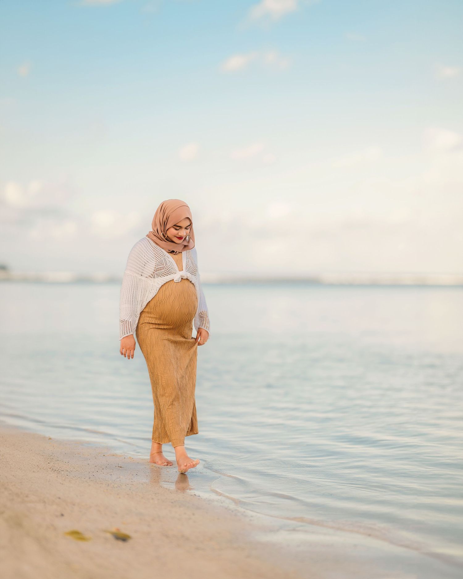 A figure in a tan dress walks along a serene beach shoreline at sunset.