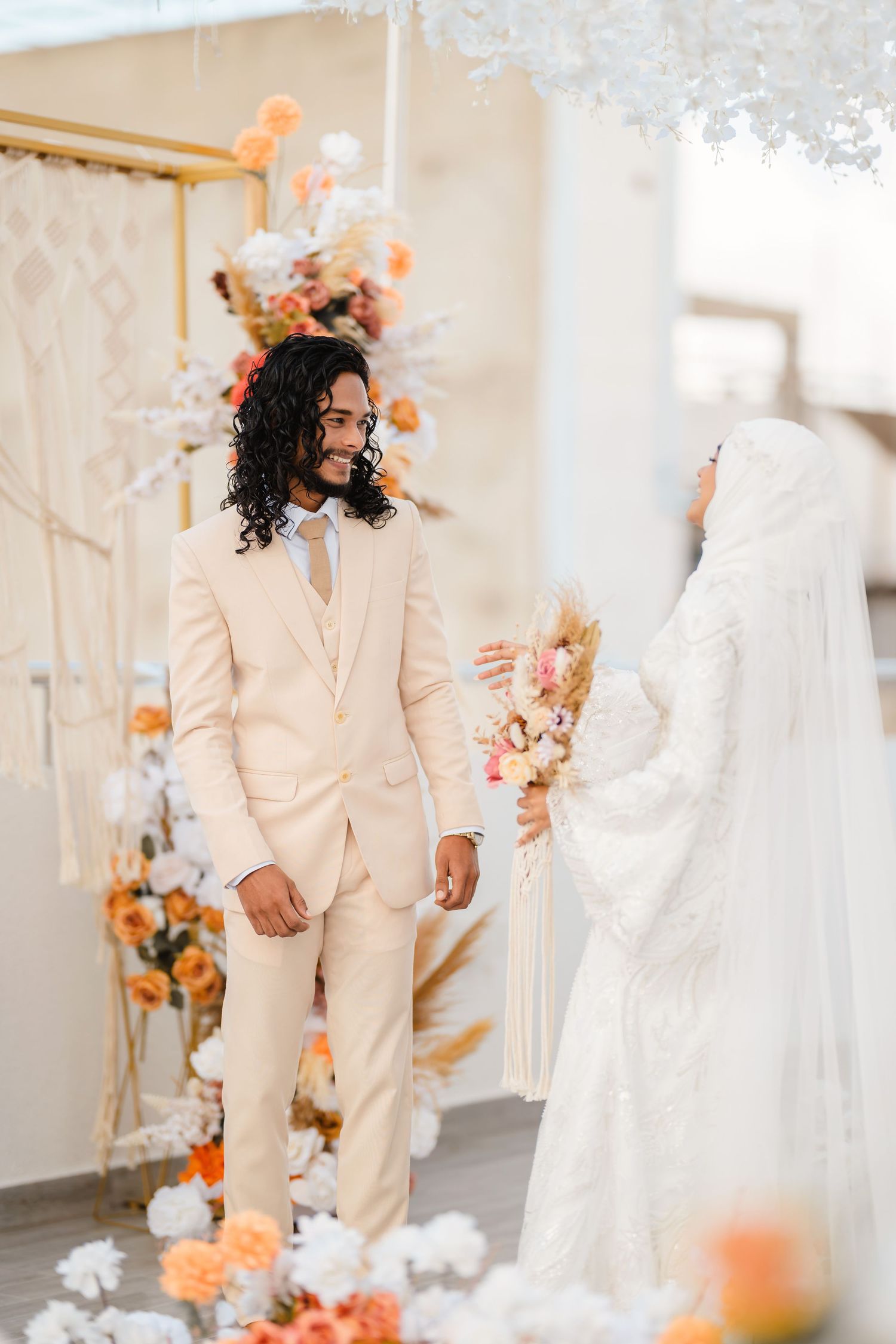 Wedding couple in white dress and cream suit pose among orange and white floral decorations.