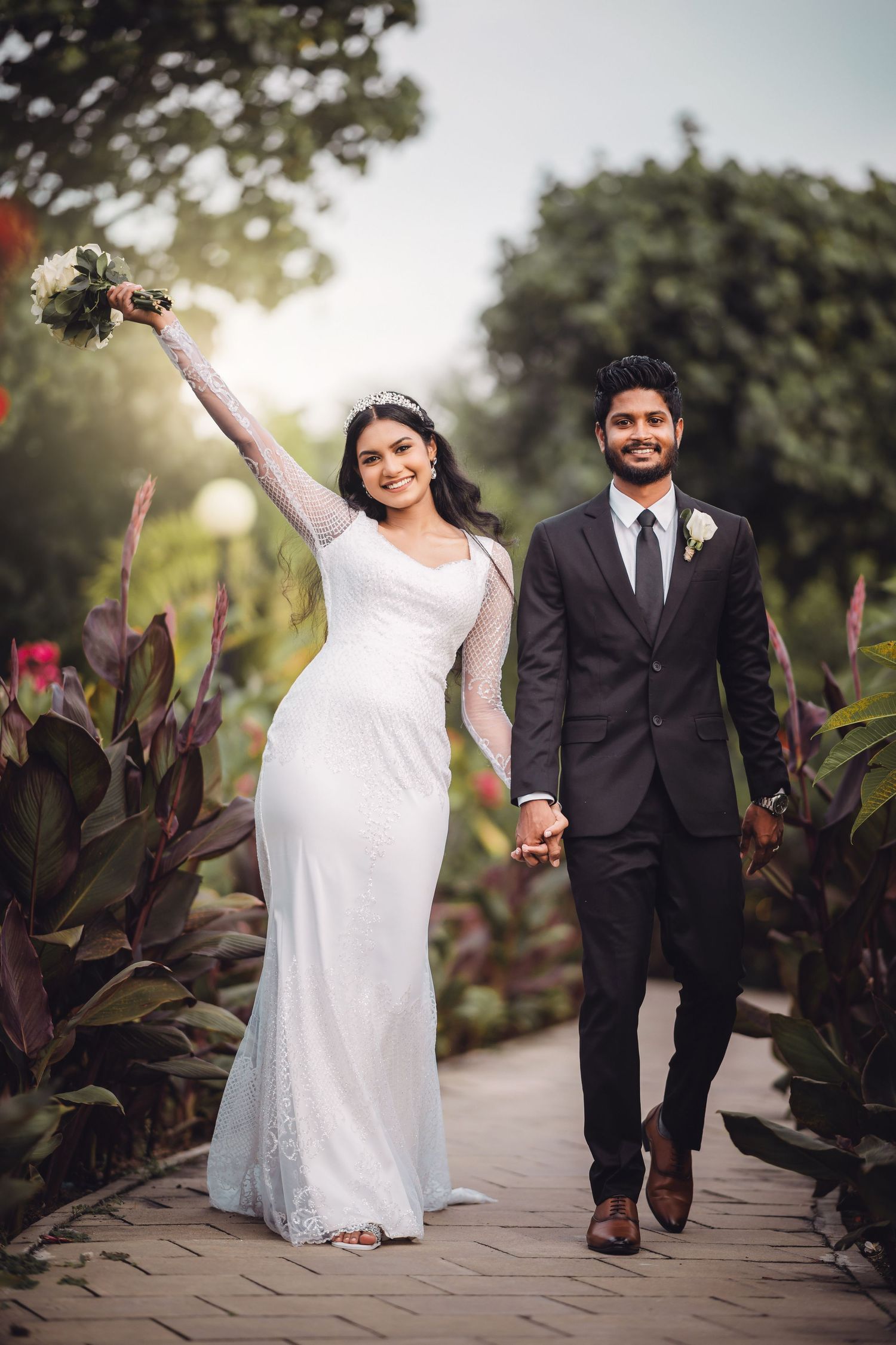 A wedding couple walks joyfully along a garden path lined with tropical plants.