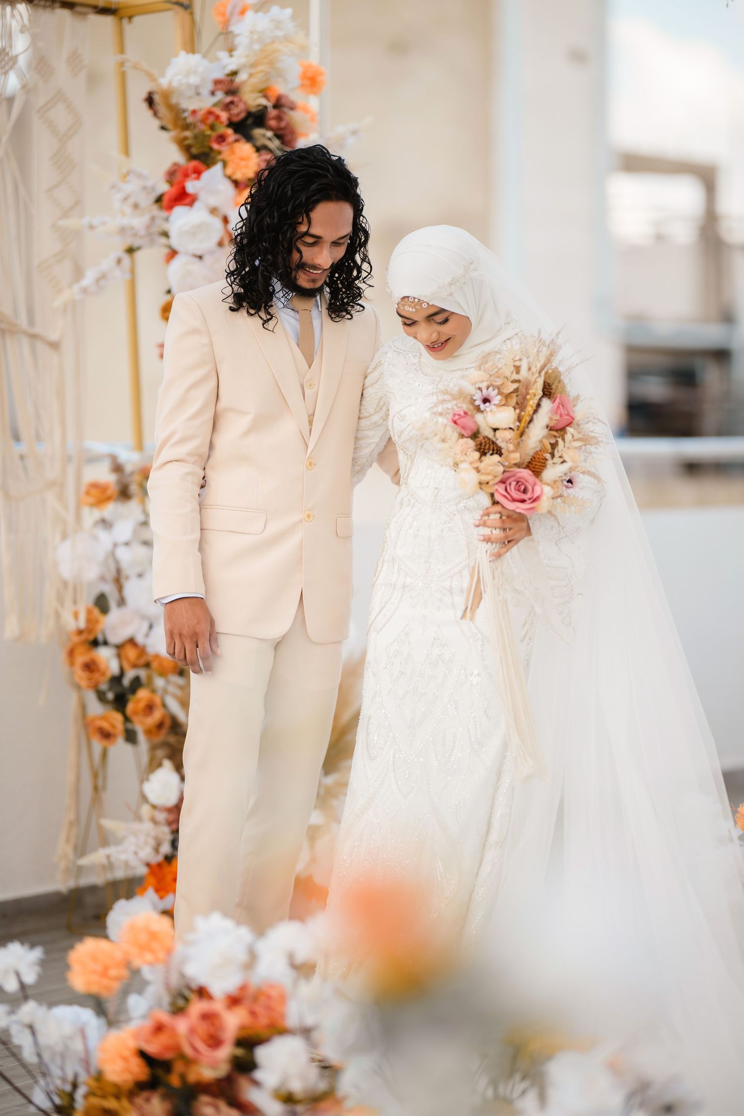 Wedding couple in white attire poses among peach and cream floral decorations.