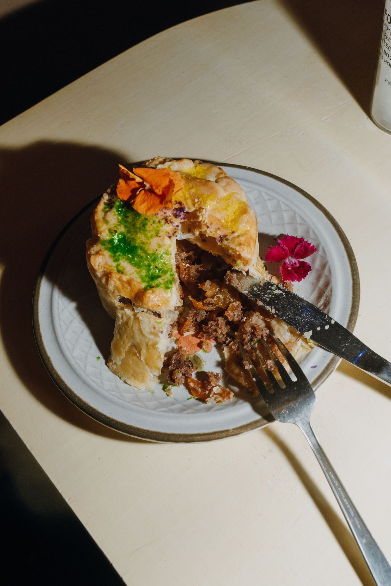 A partially eaten savory pie with ground meat filling on a white plate, garnished with edible flowers and a fork.