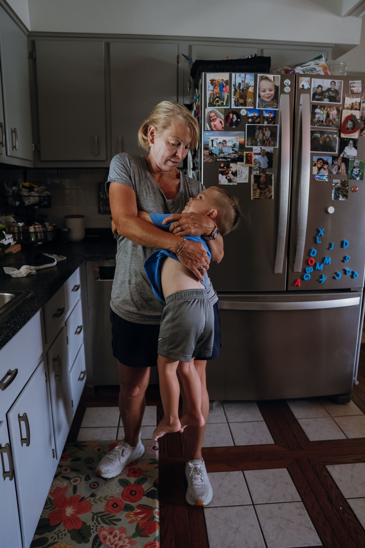 Grandmother embracing a young child in a home kitchen with photos on the refrigerator.
