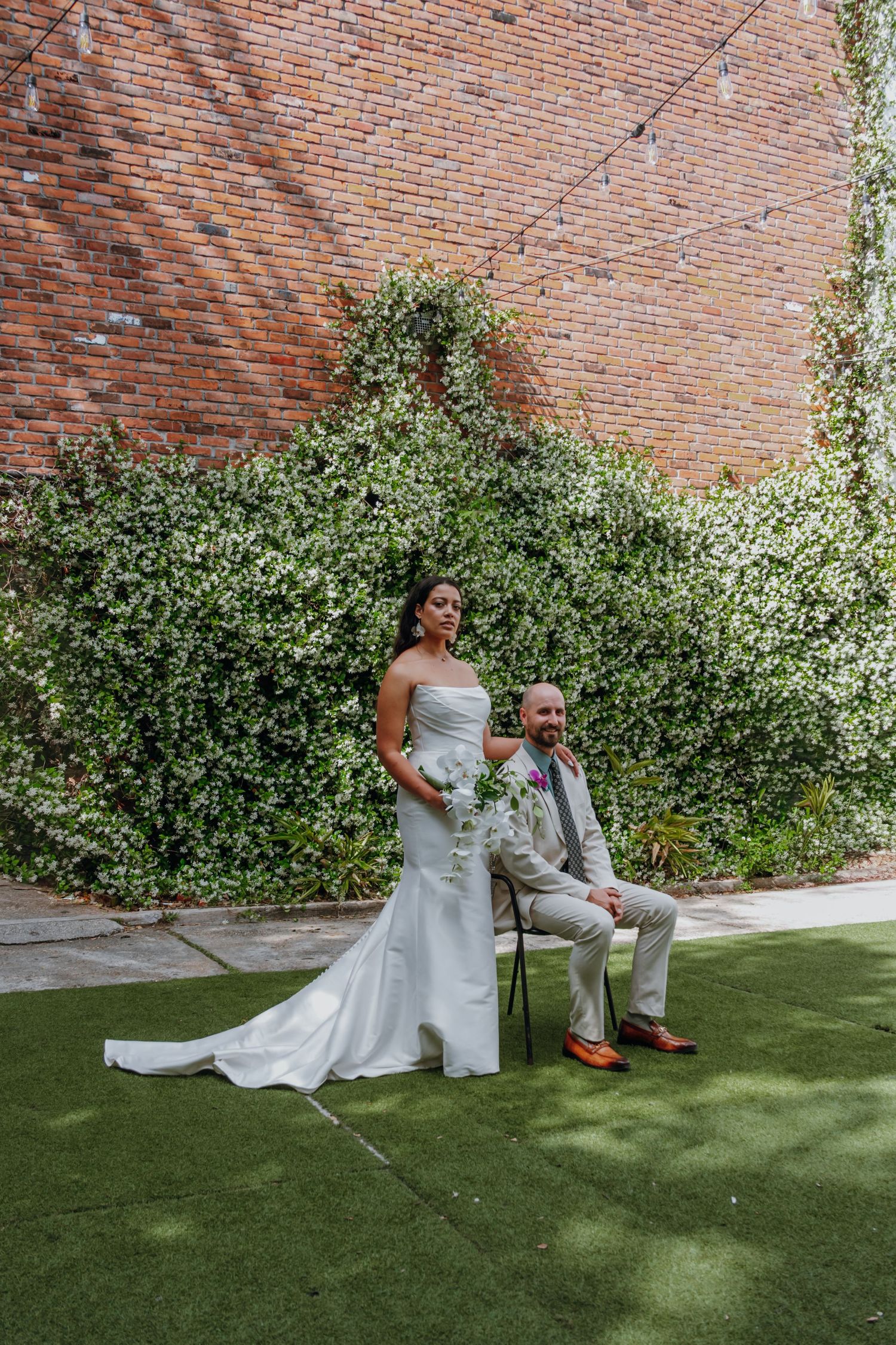 Bride and groom pose together on green lawn against ivy-covered brick wall at wedding venue.