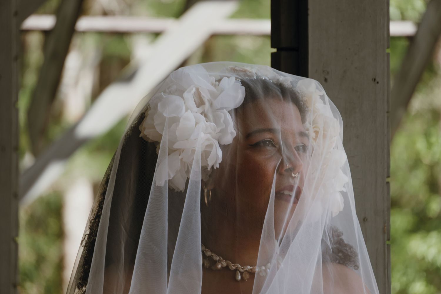 Smiling bride wearing floral white veil with flower accessories at outdoor wedding ceremony.