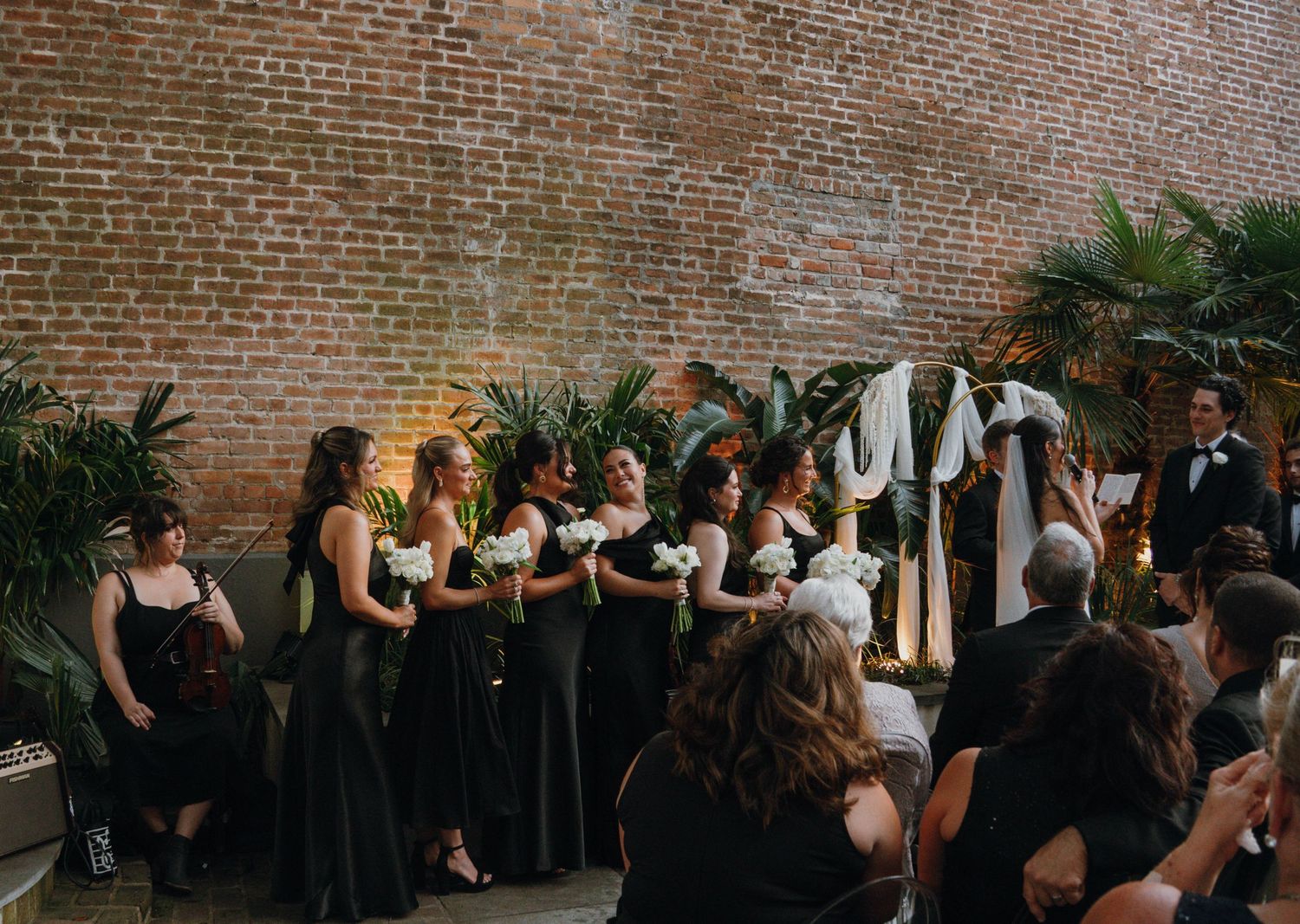 Bridesmaids in black dresses stand at altar during wedding ceremony against exposed brick wall.