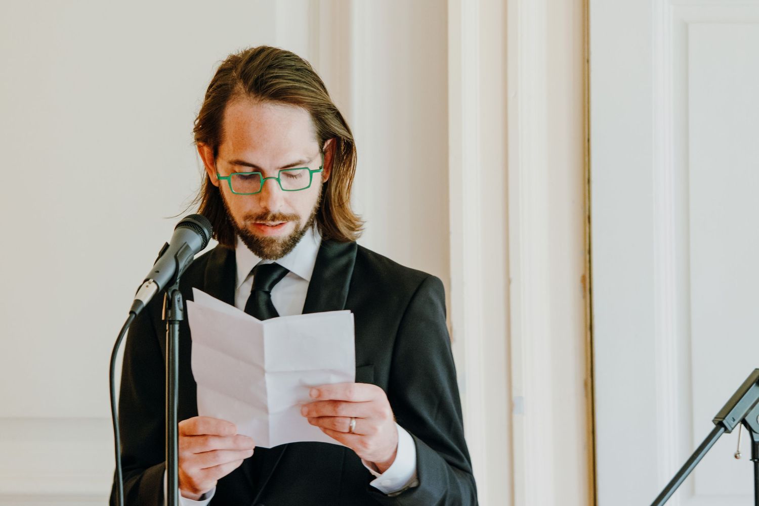 Well-dressed man with long hair and glasses reading a wedding speech at microphone.