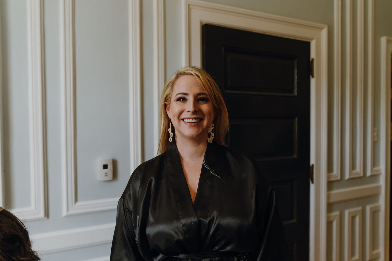 Smiling blonde bridesmaid in a black satin robe standing near a white door during wedding prep. New Orleans, Louisiana, Mississippi