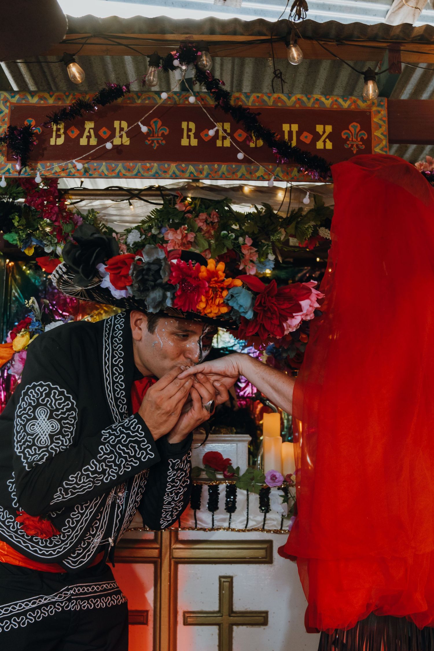 Person in traditional Mexican charro outfit kissing hand of partner in red at decorated Bar Roux altar.