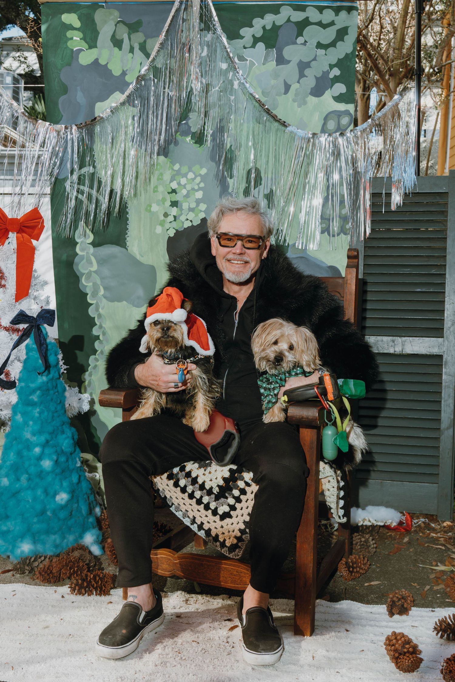 Man in black outfit sits in chair holding two small dogs in Christmas costumes, surrounded by festive holiday decorations.