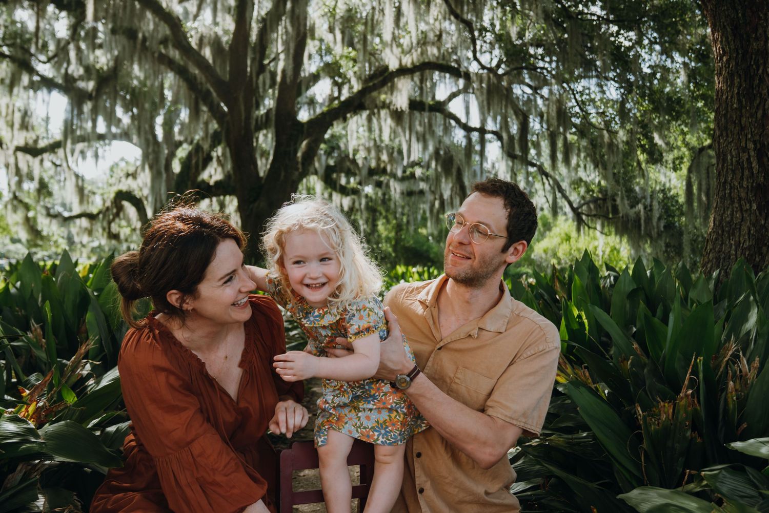 Happy family with toddler daughter smiling outdoors among lush greenery and Spanish moss-draped oak trees.
