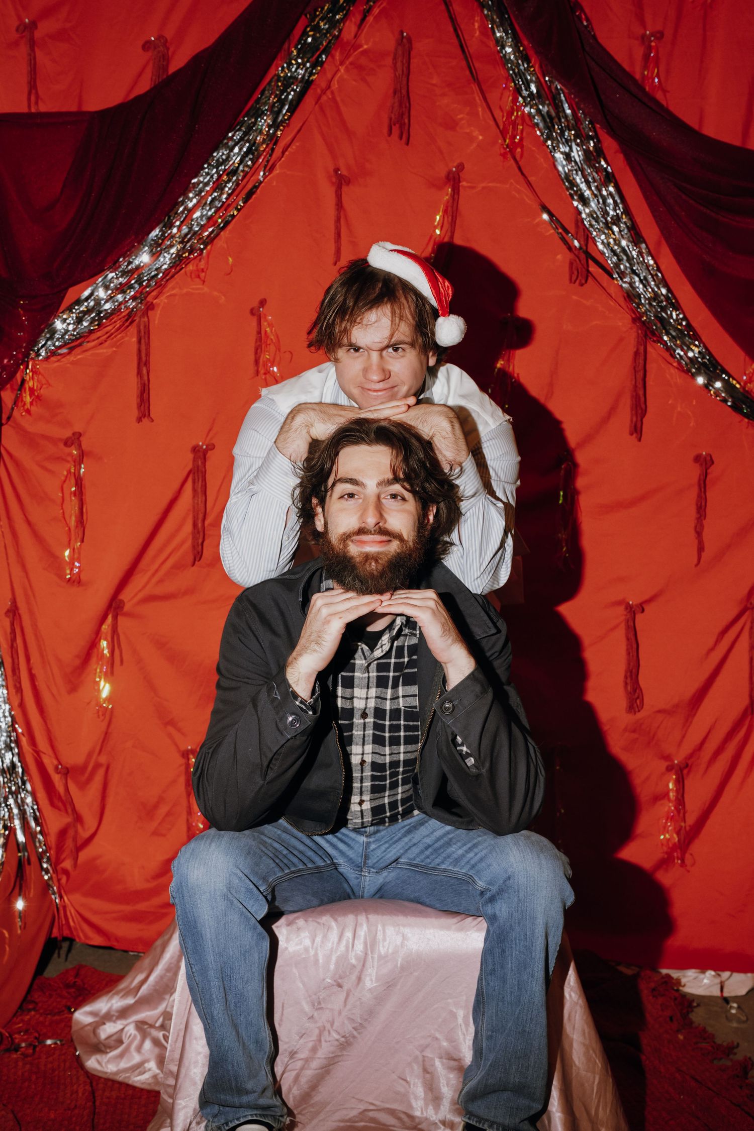 Two men pose in front of red Christmas backdrop, one wearing Santa hat, surrounded by tinsel and string lights.