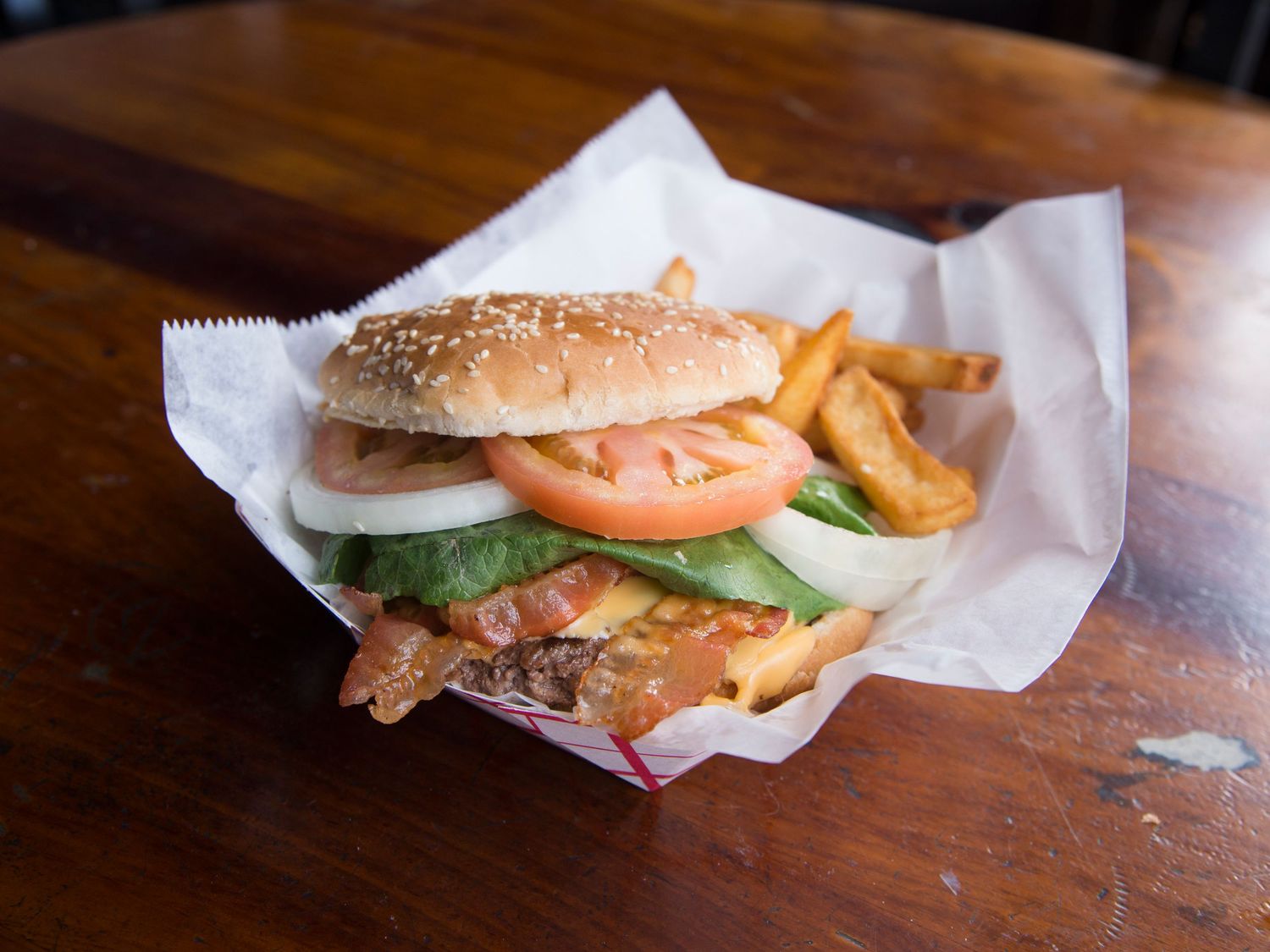 A sesame seed burger with bacon, tomato, onion, and lettuce served with thick-cut fries wrapped in white paper.