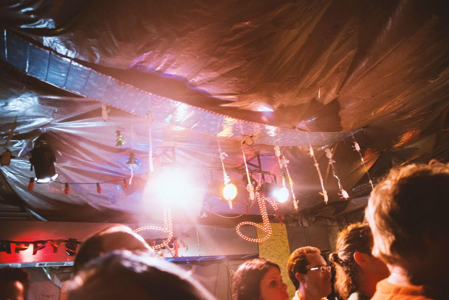 Concert crowd under plastic-draped ceiling with bright stage lights illuminating performers at an indie music venue.