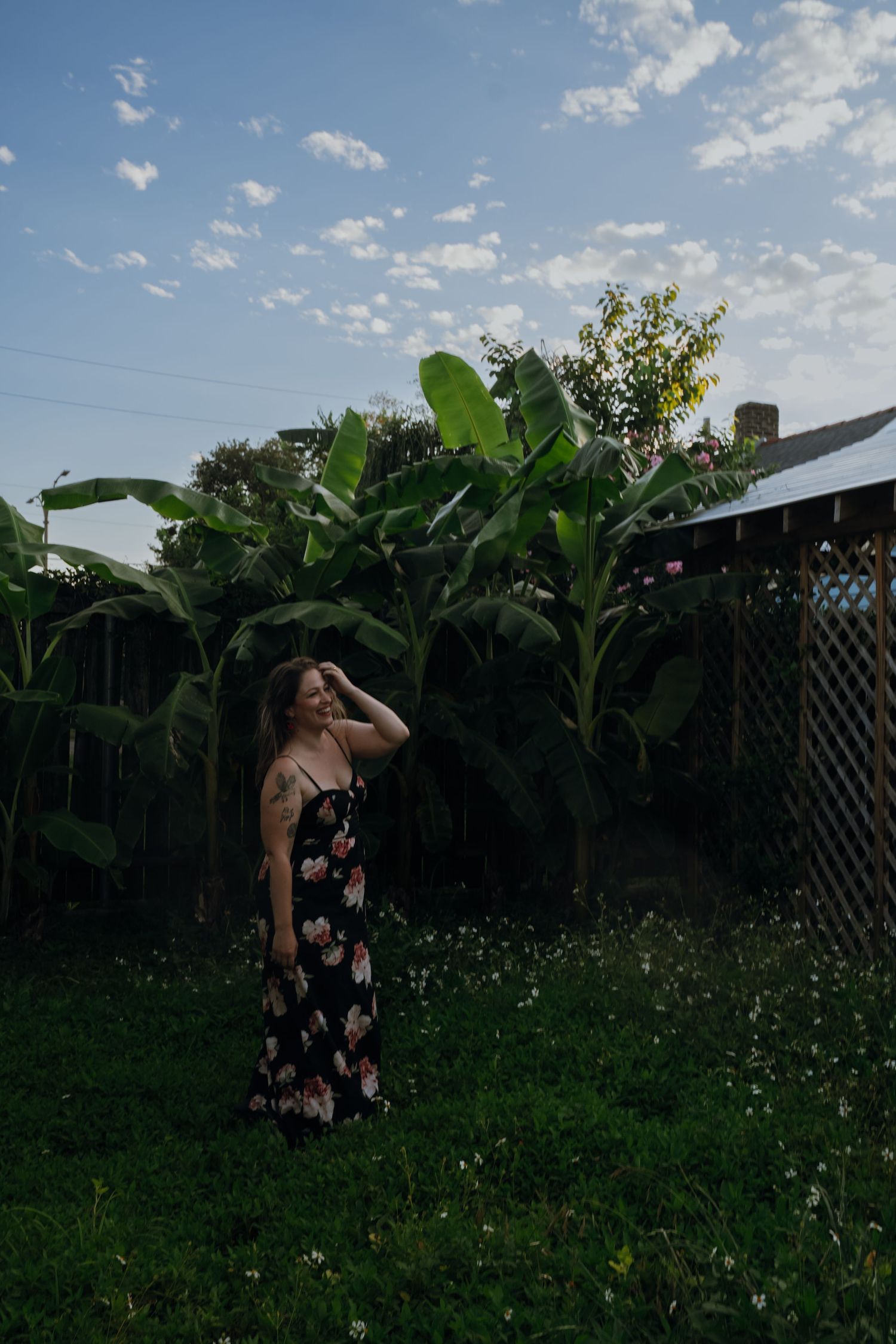 Woman in floral maxi dress standing in lush backyard garden with tropical banana plants under blue sky.