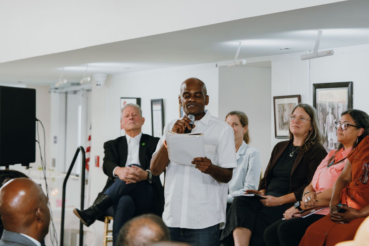 Man speaking at a community meeting, holding papers at a microphone, with seated attendees in the background.