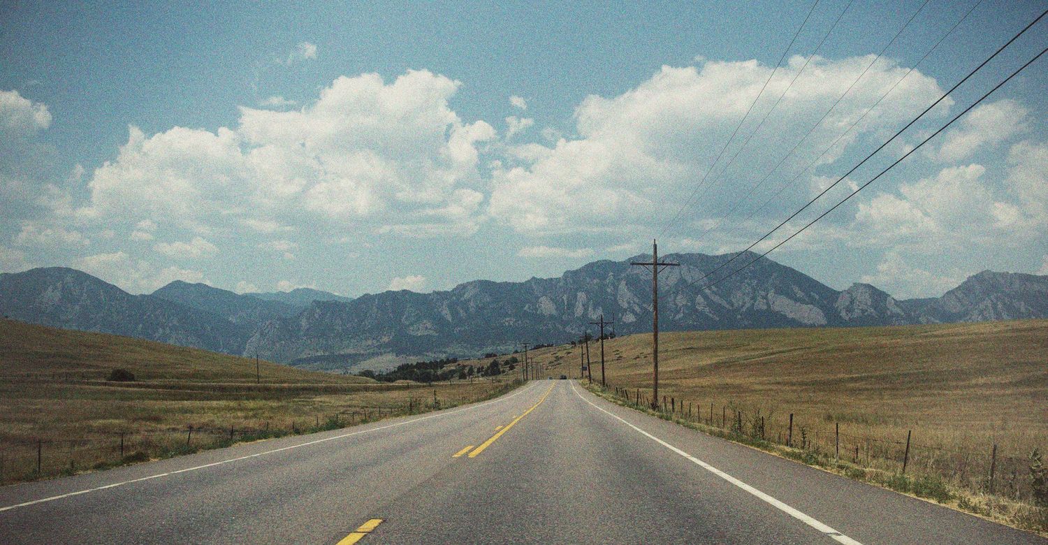 A two-lane highway stretches toward rugged mountains under a partly cloudy sky across open plains.