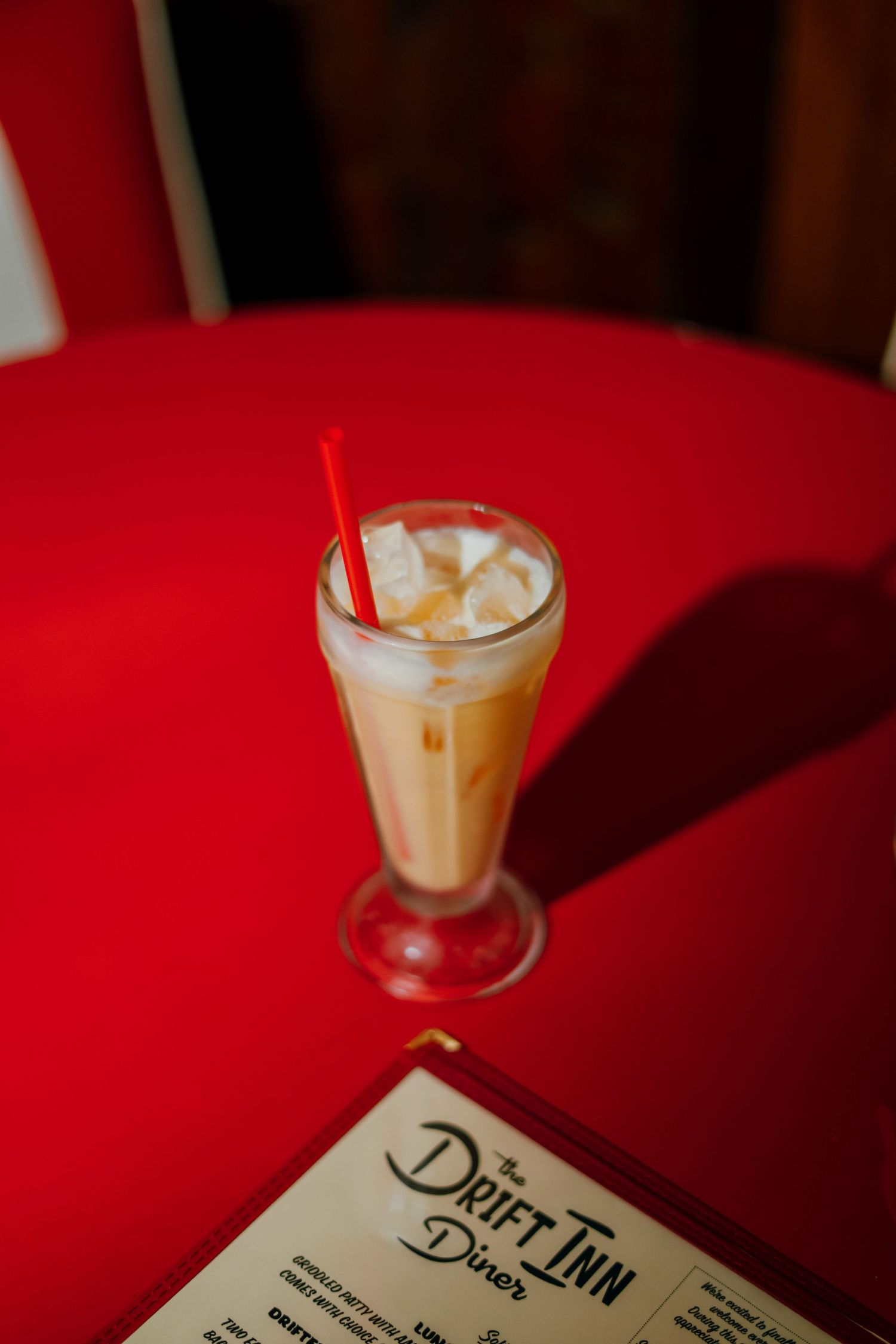 Iced coffee drink with red straw in tall glass on red table beside Drift Inn Diner menu.
